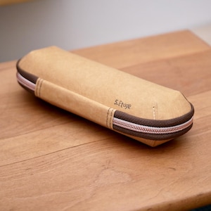 Könnte beinhalten: Ein zylindrisches, beigefarbenes Etui mit braunem Reißverschluss und Besatz. Das Etui hat einen Griff auf einer Seite und den Text "5. Etage" auf der Vorderseite. Der Reißverschluss hat ein hellrosa und weiß gestreiftes Design.