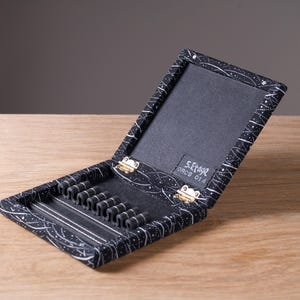 Può includere: Un astuccio rettangolare nero con un motivo a linee astratte bianche, dotato di coperchio incernierato e interno nero con sei passanti elastici. Una piccola etichetta all'interno del coperchio riporta il testo 5.Etage ORCS 014 in bianco.
