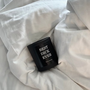 Op de afbeelding: Een zwarte rechthoekige kaars met de tekst "BEST DICK EVER" in het wit, liggend op een wit laken. De kaars heeft een matte afwerking en een etiket met de merknaam "Veyre concept".
