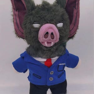 May include: A plush bat doll with a green furry head, large pink ears, and a pink snout. It wears a blue blazer, a white shirt, a red tie, black pants, and black shoes. The doll has white eyes and a small mouth.