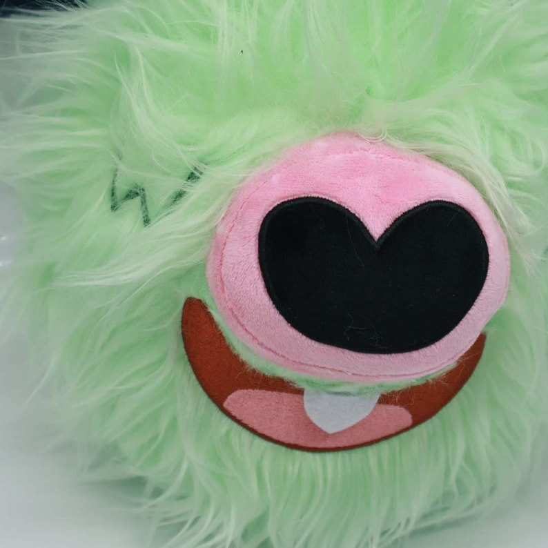 Puede incluir: Una cara de monstruo de peluche con pelaje lanudo verde claro. El monstruo tiene una nariz rosa con una boca negra en forma de coraz&oacute;n, una boca marr&oacute;n y un diente blanco. Un garabato verde es visible en la piel.