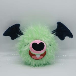 Puede incluir: Un peluche de monstruo con un cuerpo redondo verde claro cubierto de pelaje hirsuto. Tiene una nariz rosa con una fosa nasal negra en forma de coraz&oacute;n, una boca roja sonriente y dos alas azul oscuro.