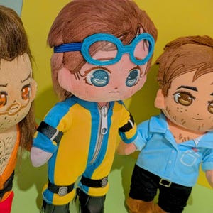 May include: Three plush dolls with embroidered facial features and brown hair. The left doll wears an orange top. The center doll wears a yellow and blue jumpsuit with blue goggles. The right doll wears a light blue shirt and black pants.