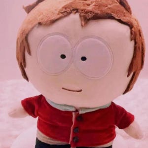 Könnte beinhalten: Plüschpuppe einer Zeichentrickfigur mit rundem Kopf, großen Augen und braunen Haaren. Die Puppe trägt eine rote Jacke mit dunklen Knöpfen, einen hellblauen Kragen und eine schwarze Hose. Die Puppe steht auf einer weißen Oberfläche.