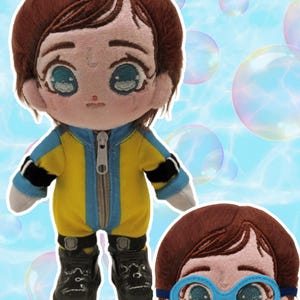 May include: A soft toy doll with brown hair and large eyes. The doll is dressed in a yellow and blue jumpsuit with a zip and black boots. A second doll head with blue goggles is also visible. The background has a light blue gradient with floating bubbles.