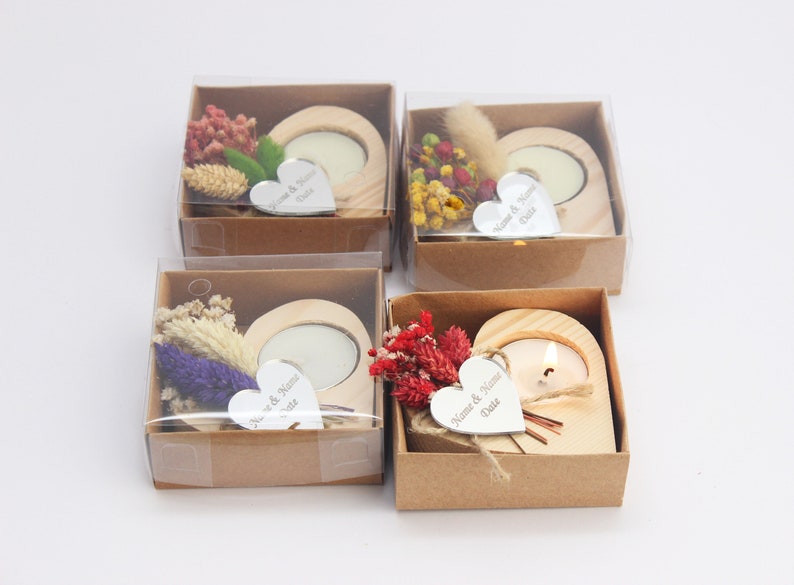 May include: Four small, brown cardboard boxes with clear plastic lids. Each box contains a heart-shaped wooden candle holder with a small white tea light candle. The candle holders are decorated with dried flowers and a small silver heart-shaped tag with the text "Name & Name Date" engraved on it.