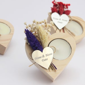May include: Three wooden heart-shaped candle holders with a small tea light candle in each. Each holder has a small silver heart tag with the text "Name & Name Date" engraved on it. The holders are decorated with dried flowers.