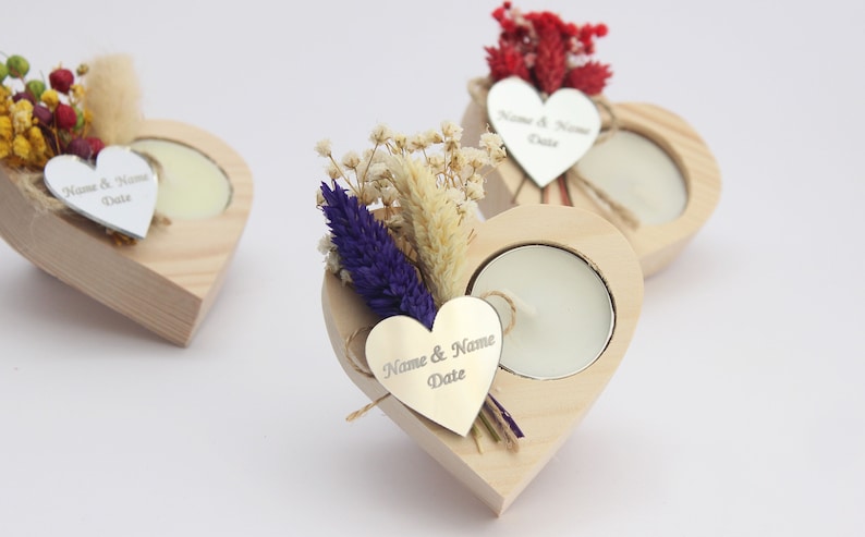 May include: Three wooden heart-shaped candle holders with a small tea light candle in each. Each holder has a small silver heart tag with the text "Name & Name Date" engraved on it. The holders are decorated with dried flowers.