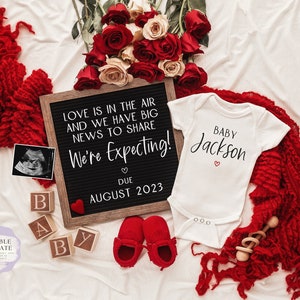 May include: A pregnancy announcement with a letter board that reads "Love is in the air and we have big news to share. We're expecting! Due August 2023". There is a white onesie with the text "Baby Jackson" and a red heart. There are red roses, a red blanket, and wooden blocks that spell "Baby".