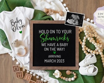 May include: A green and white baby announcement with a letter board that reads "Hold on to your Shamrocks We have a baby on the way! Arriving March 2023". The announcement also includes a baby onesie, a pair of baby shoes, a wooden bead garland, and a wooden rattle.