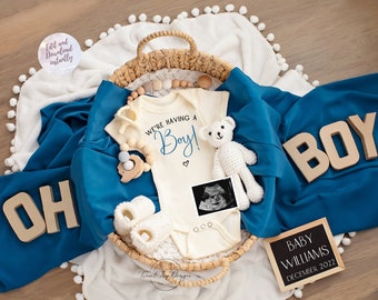 Puede incluir: Un body blanco para bebé con el texto "We're having a Boy!" en letras azules. El body está en una cesta de mimbre con una manta azul, un osito de peluche, un sonajero de madera y una foto de ecografía. La cesta está rodeada de una manta blanca con pompones blancos. Hay letras de madera que deletrean "OH BOY" en la manta.