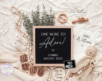 Könnte beinhalten: Eine Babyankündigung mit einer schwarzen Filz-Buchstabentafel mit der Aufschrift "ONE MORE TO Adore!" und "COMING AUGUST 2023". Das Bild enthält Babykleidung, Holzblöcke, einen Plüschbären und eine Holzrassel.