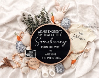 May include: A black letter board with the text "We are excited to say that a little Somebunny is on the way! Arriving December 2023".  The board is surrounded by a variety of baby items, including a white onesie with the text "Baby Johnson", a stuffed bunny, and a wooden bead garland.