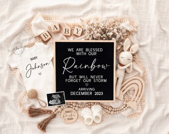 May include: A black letter board with the text "We are blessed with our Rainbow but will never forget our storm arriving December 2023". The board is surrounded by baby items, including a white onesie with the text "Baby Johnson", a wooden rainbow, a crocheted bunny, and a wooden sign that says "Sweet Rainbow Baby".