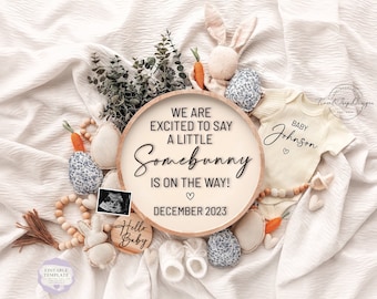 May include: A wooden round sign with the text "We are excited to say a little Somebunny is on the way!" and "December 2023". The sign is surrounded by baby items, including a onesie, a bunny, and a pacifier.
