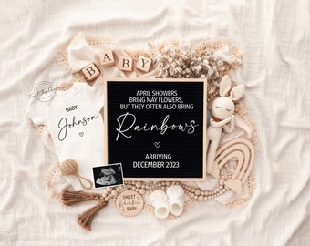 May include: A black letter board with the text "April showers bring may flowers, but they often also bring Rainbows arriving December 2023". The board is surrounded by baby items, including a white onesie with the text "Baby Johnson", a wooden rainbow, a crocheted bunny, and a wooden sign that says "Sweet Rainbow Baby".