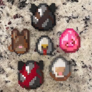 May include: A collection of six pixel art figures crafted from small plastic beads. The set includes a brown animal head, a bright pink egg, a red and black patterned egg, a dark grey egg, a white and grey egg, and a small white and grey egg.
