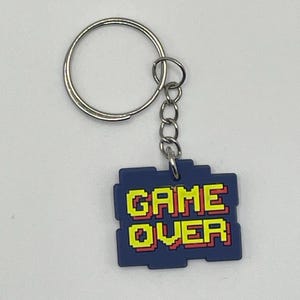 May include: A rectangular blue keychain featuring the pixelated text GAME OVER in yellow with a red outline. The charm is attached to a silver-coloured metal split ring by a short chain. The design has a blocky, stepped edge reminiscent of retro video game graphics.