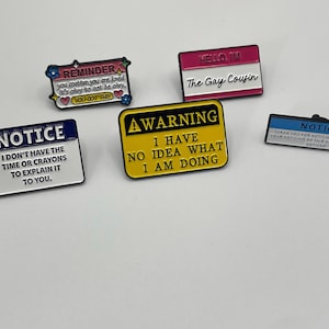 May include: Five rectangular enamel pins with text. One says REMINDER YOU MATTER, one says HELLO I'M The Gay Cousin, one says NOTICE I DON'T HAVE THE TIME OR CRAYONS TO EXPLAIN IT TO YOU, one says WARNING I HAVE NO IDEA WHAT I AM DOING, and one says NOTICE.