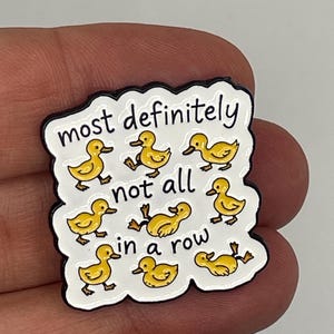 May include: A white enamel pin with a black border featuring eight yellow ducklings in various positions and the black text "most definitely not all in a row" written across the centre of the pin in a casual, handwritten style font.