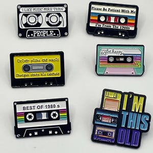 May include: A collection of six enamel pins featuring retro cassette tape designs. The pins display various phrases including I LIKE MUSIC MORE THAN PEOPLE, Please Be Patient With Me I'm From The 1900s, Driver picks the music Shotgun shuts his cakehole, Be happy, BEST OF 1980 s, and I'M THIS OLD.