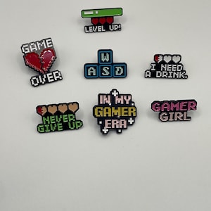 May include: A collection of seven enamel pins featuring pixel art designs. The pins include text such as GAME OVER, LEVEL UP!, I NEED A DRINK., NEVER GIVE UP, IN MY GAMER ERA, and GAMER GIRL, alongside icons like hearts, a keyboard layout, and a health bar.