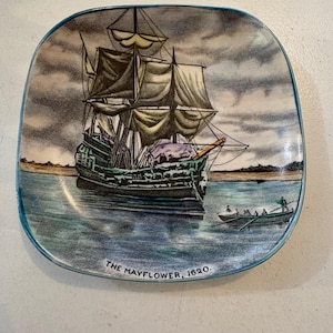 May include: A decorative plate featuring a detailed illustration of the Mayflower ship sailing on water under a cloudy sky. The plate has a blue border and the text "THE MAYFLOWER, 1620."