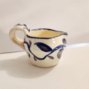 May include: A small cream pitcher with a cream-colored base and a hand-painted blue vine pattern featuring dark purple leaves. The ceramic vessel has a rounded body, a curved handle on the side, and a small spout for pouring liquids.