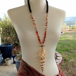 May include: A long necklace with a large seashell pendant. The necklace is made of black beads, orange beads, and white shell beads. The seashell is a spiral shape and is a light brown color.
