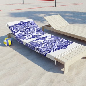 Puede incluir: Una toalla de playa blanca con un patrón geométrico azul y blanco. La toalla está doblada y colocada en una tumbona de madera.