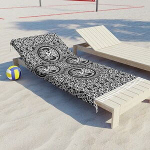 Puede incluir: Toalla de playa negra y blanca con un diseño de árbol de la vida. La toalla está colocada en una tumbona de madera en una playa de arena.