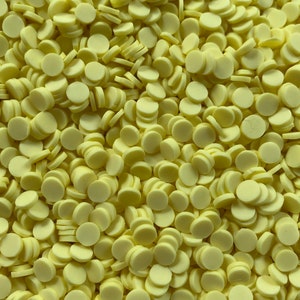 May include: A close-up shot of a pile of small, pale yellow, circular objects. The objects are disc-shaped with a slightly raised edge, resembling small buttons or candies. The items are scattered, creating a textured, dense pattern.