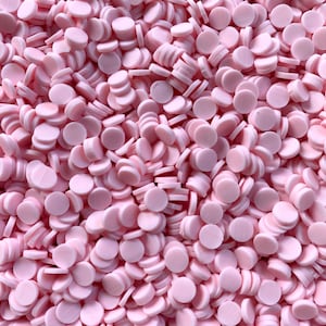 May include: A close-up shot of a pile of small, light pink, circular polymer clay pieces. The pieces are stacked and scattered, creating a textured surface. The clay pieces appear to be uniform in size and shape.