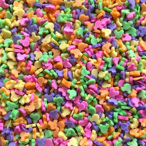 May include: A close-up of a large quantity of colorful sprinkles shaped like Mickey Mouse heads. The sprinkles are in shades of yellow, orange, green, pink, and purple.