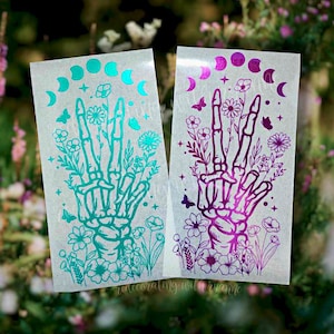 May include: Two sticker sheets with a skeleton hand making a peace sign surrounded by flowers and a crescent moon phase design. The sticker on the left is teal and the sticker on the right is purple.