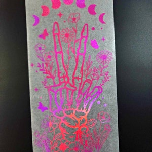 May include: A holographic sticker with a pink and purple gradient. The sticker features a hand with three fingers raised, surrounded by flowers and a crescent moon.