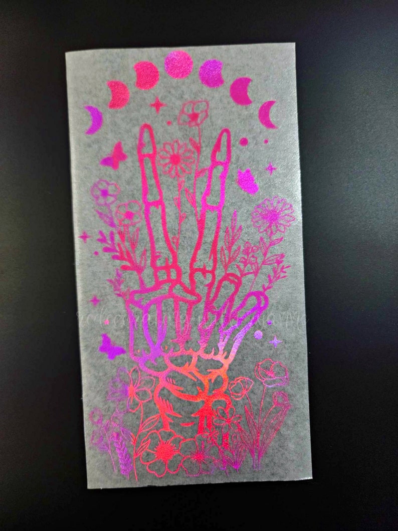 May include: A holographic sticker with a pink and purple gradient. The sticker features a hand with three fingers raised, surrounded by flowers and a crescent moon.