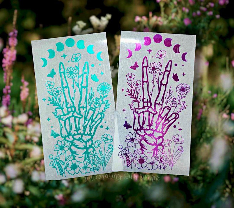 May include: Two sticker sheets with a skeleton hand making a peace sign surrounded by flowers and a crescent moon phase design. The sticker on the left is teal and the sticker on the right is purple.