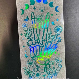 May include: A holographic sticker featuring a skeletal hand making a peace sign surrounded by flowers and a crescent moon design.