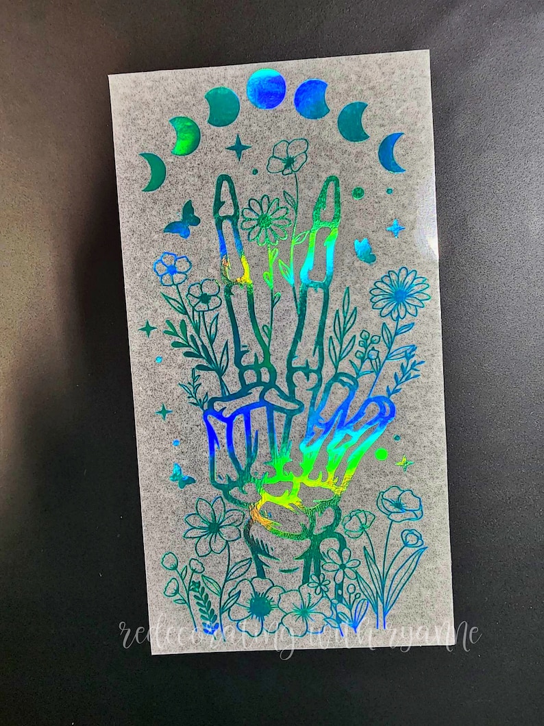 May include: A holographic sticker featuring a skeletal hand making a peace sign surrounded by flowers and a crescent moon design.