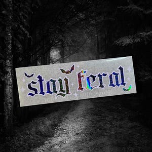 May include: A white sticker with a holographic finish featuring the text "Stay Feral" in a gothic font. The sticker is decorated with small stars and bat silhouettes.