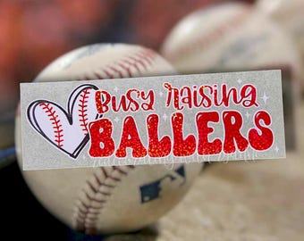 Puede incluir: Un balón de béisbol blanco con costuras rojas y un corazón blanco y rojo con un diseño de béisbol. Una pegatina blanca con el texto "Busy raising BALLERS" en letras rojas de purpurina.