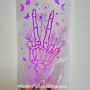 May include: A pink iridescent vinyl decal featuring a skeleton hand making a peace sign with flowers and moons around it. The decal has the text "@RedecoratewithRyanne" at the bottom.