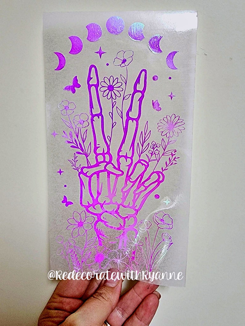 May include: A pink iridescent vinyl decal featuring a skeleton hand making a peace sign with flowers and moons around it. The decal has the text "@RedecoratewithRyanne" at the bottom.