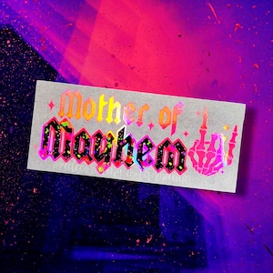 May include: A holographic sticker with the text "Mother of Mayhem" in a gothic font with a pink and yellow color scheme. A skeleton hand with a middle finger is on the right side of the sticker.
