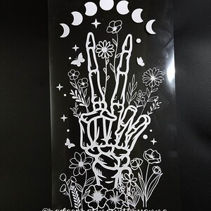May include: White vinyl decal featuring a skeleton hand with flowers and crescent moons. The decal includes the text "@redecoratingwithryanne".