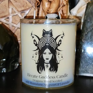 May include: A white candle in a glass jar with a black and white label that reads "Hecate Goddess Candle Dragons Blood Scented with Moonstone, Mugwort & Lavender". The label features a black and white illustration of a woman with three faces, a wolf, and a crescent moon.
