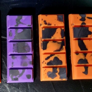 May include: Five wax melts in a variety of colors and patterns. The melts are in a rectangular shape with six sections. The colors are orange, black, purple, and gold. The patterns are swirls and dots.