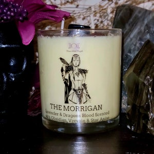 May include: A candle with a black and white illustration of a woman holding a raven. The candle is labeled "The Morrigan" and has a description of "Lavender & Dragons Blood Scented with Obsidian, Vervain & Star Anise".