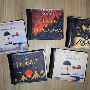 May include: Five black wallets with book cover designs. Titles include "Queen of Shadows", "The Hobbit", "Zodiac Academy", and "the things we leave unfinished". The wallets are rectangular and made of a leather-like material.